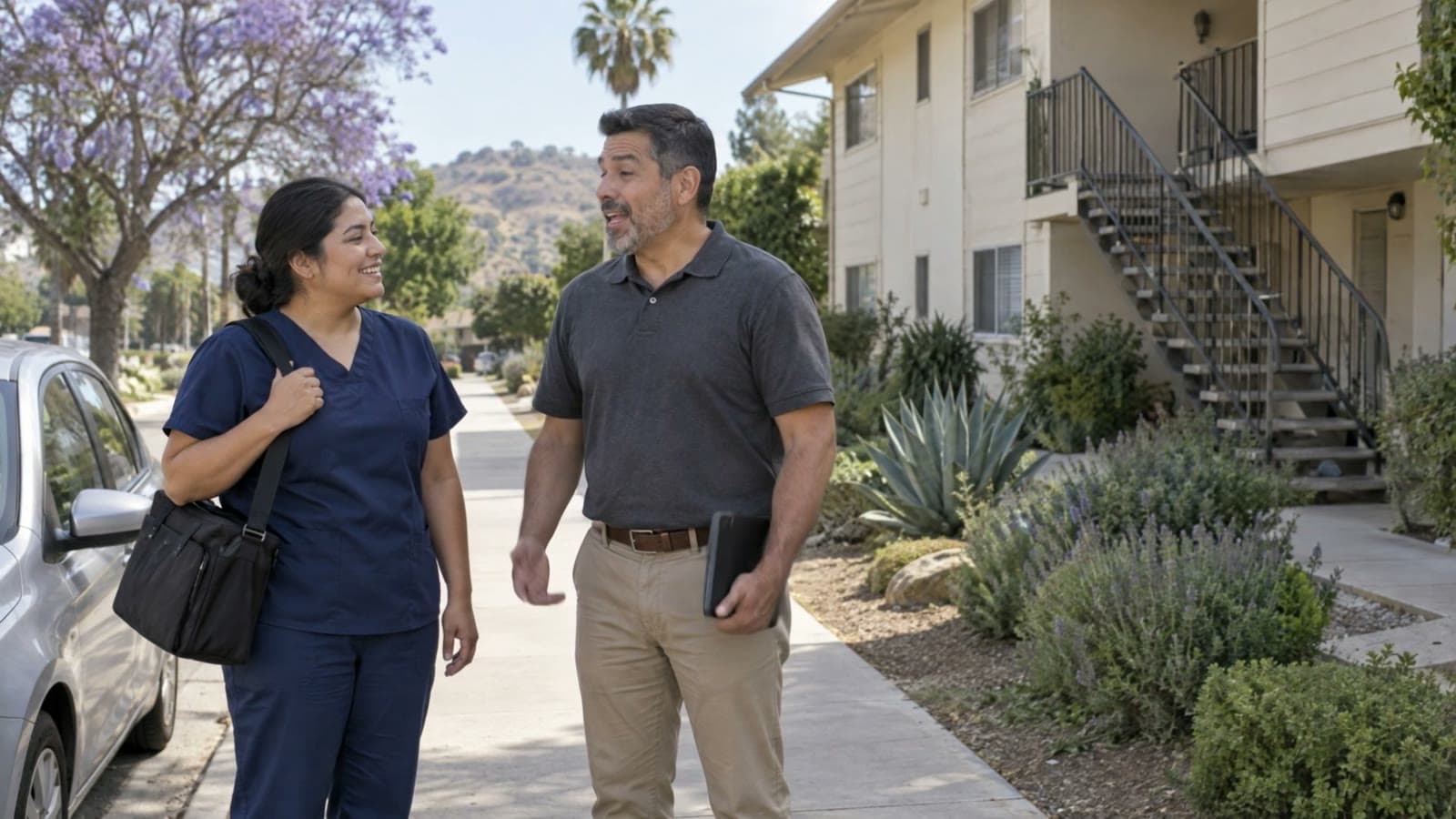 A woman in navy scrubs with a shoulder bag and a man in a gray polo holding a tablet laugh together on a sidewalk beside a parked car, in front of a two-story apartment building.