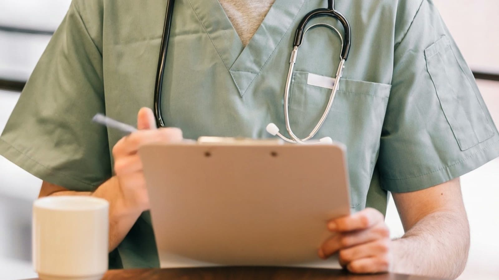 A clinician in green scrubs reads a clipboard at a wooden table beside a white mug.