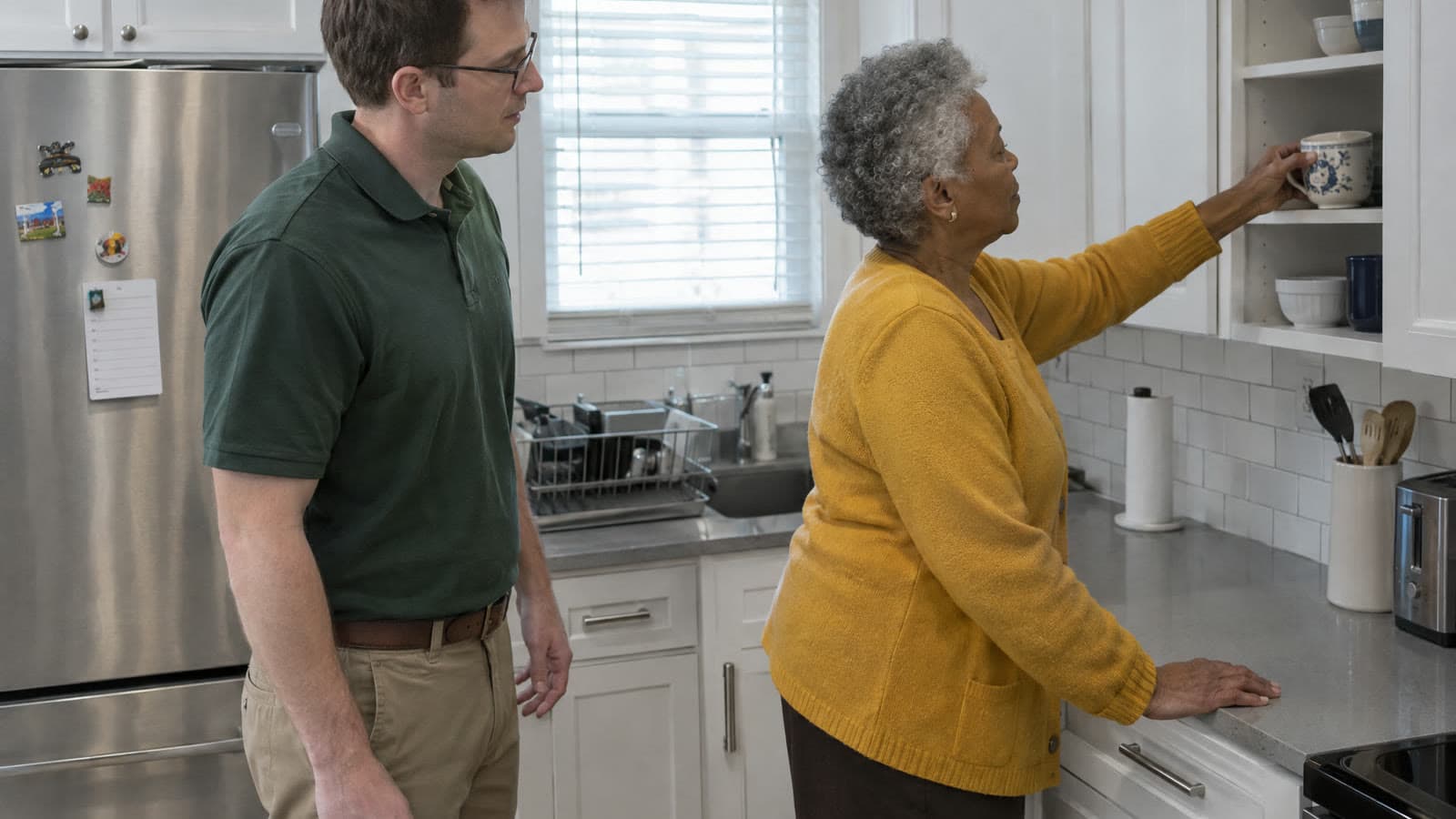 An older woman in a mustard cardigan sets a mug on a kitchen shelf while a man in a green polo stands beside her, watching.